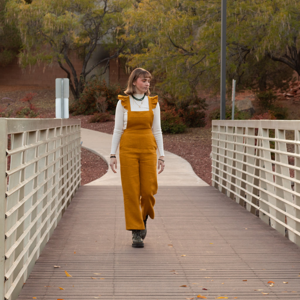 Person wearing mustard yellow overalls walking on a wooden bridge with trees in the background