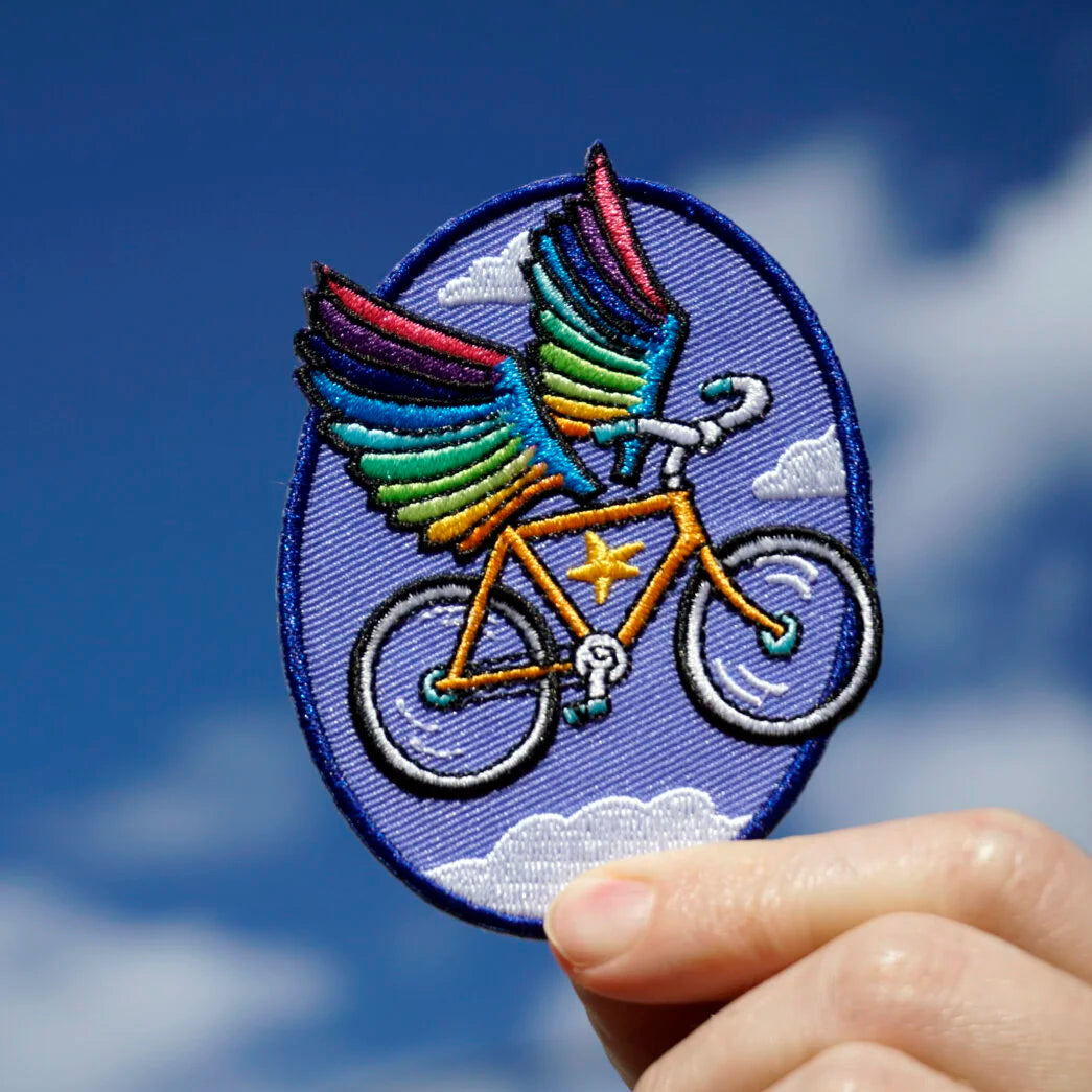Embroidered patch of a bicycle with rainbow wings held against a blue sky.