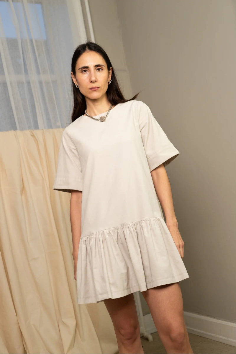 Person wearing a beige dress standing in a room with curtains and a neutral wall.