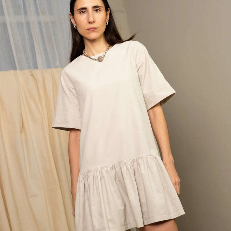 Person wearing a beige dress standing in a room with curtains and a neutral wall.