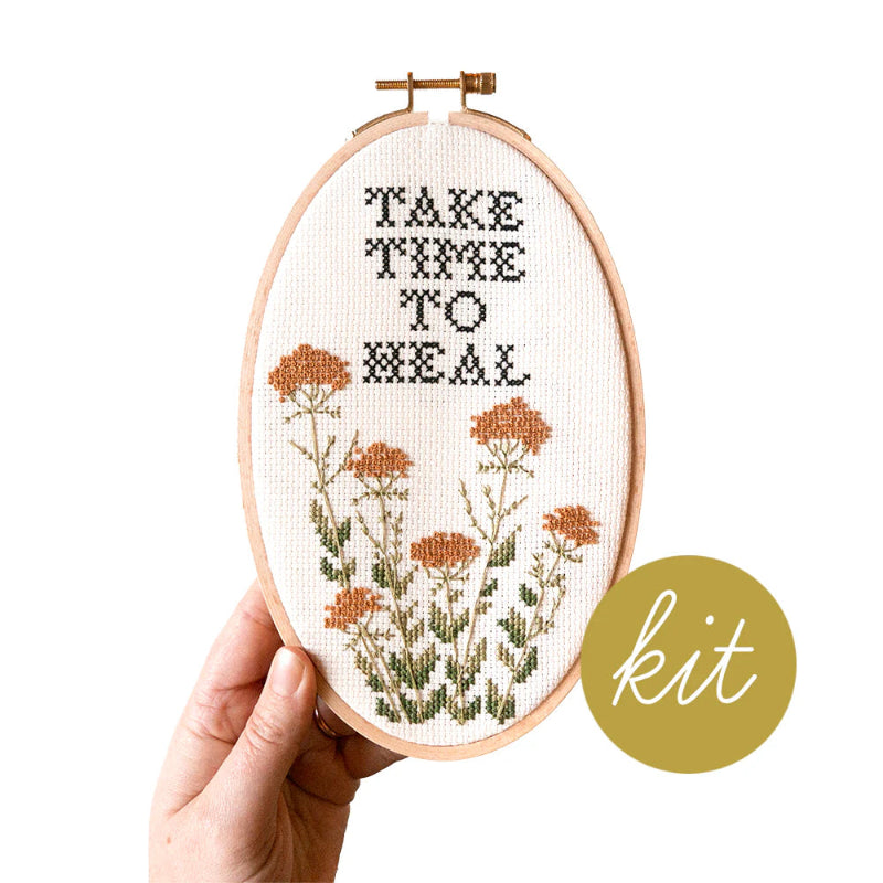 Embroidery hoop with cross-stitched design and text 'Take Time to Heal' held by a hand on a white background.