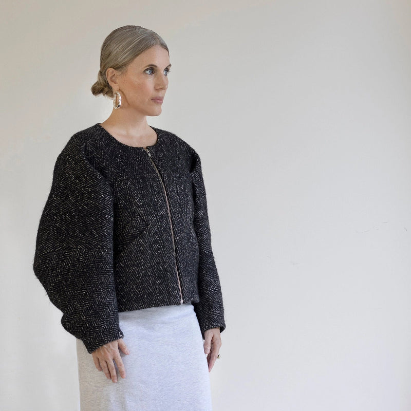 person wearing a black textured jacket and white pants against a plain background