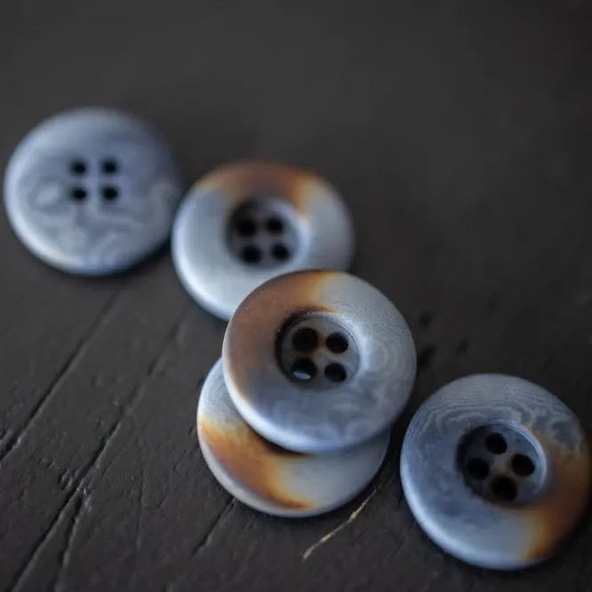 Five round decorative buttons with a marbled light blue-gray and white pattern and brown burnished edges, arranged on a dark textured surface.