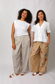Two women modeling loose, wide-leg pants in a studio setting. The woman on the left wears light beige textured wide-leg pants with a white sleeveless top, and the woman on the right wears tan wide-leg pants with colorful butterfly prints and a white short-sleeve blouse.