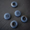 Five round buttons with a textured, metallic blue-gray finish arranged on a dark, rustic wooden surface.