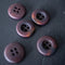 Five round four-hole buttons with a metallic bronze and purple iridescent finish, arranged on a dark textured surface.